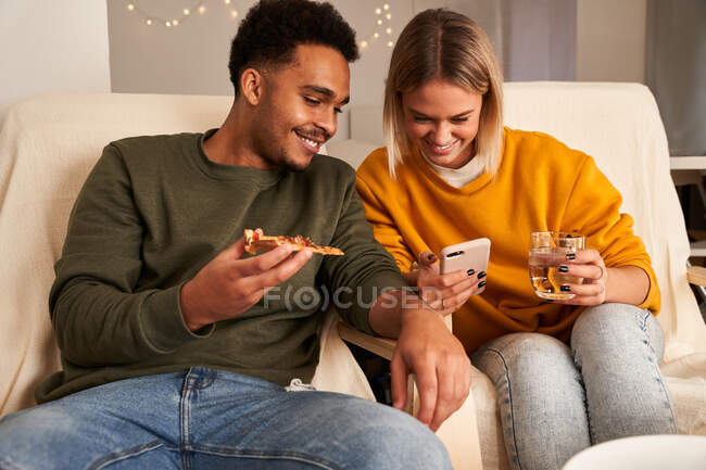 Pareja multirracial contenido sentado en la sala de estar mientras come pizza y ver videos divertidos en el teléfono inteligente en fin de semana - foto de stock