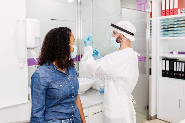 Médico en guantes de látex y protector facial que rellena la jeringa del frasco con la vacuna que se prepara para vacunar a una paciente afroamericana irreconocible en la clínica durante el brote de coronavirus - foto de stock