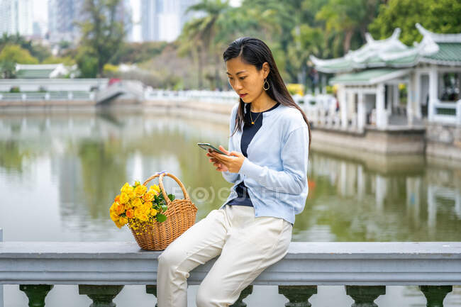 Belle asiatique fille portrait dans un parc alors qu'elle est assise et regarde son téléphone portable à côté de panier en osier avec des fleurs jaunes. — Photo de stock