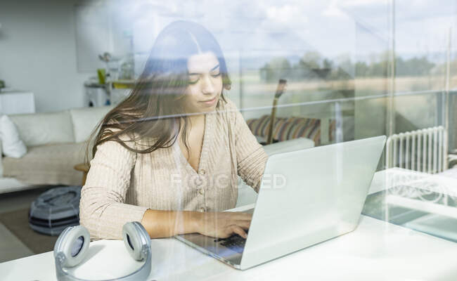 Through glass view of cheerful young female browsing internet on netbook at table with headset in apartment — Stock Photo
