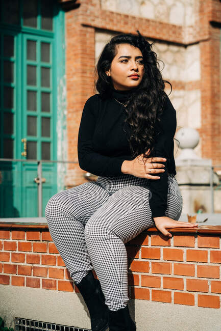 Confiante plus tamanho feminino no casaco da moda sentado na borda de pedra perto de edifício de tijolo velho e olhando para longe — Fotografia de Stock