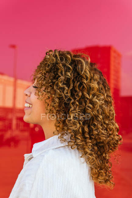 Vue latérale de la femme afro-américaine ravie avec coiffure bouclée debout sur fond rouge en studio regardant loin — Photo de stock