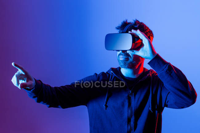 Varón barbudo irreconocible con capucha y auriculares modernos que exploran la realidad virtual con luz de neón - foto de stock