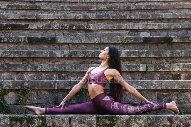 Seitenansicht einer flexiblen Frau in aktiver Kleidung, die in Hanumanasana in Steintreppen sitzt und ihren Körper dehnt, während sie Yoga praktiziert — Stockfoto