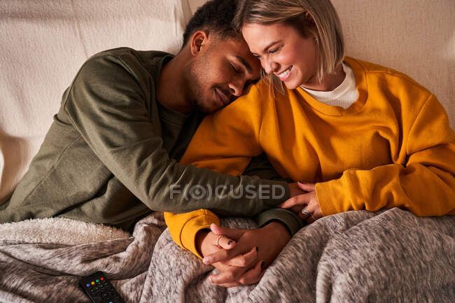 Alto ángulo de amorosa pareja multiétnica relajándose en el sofá bajo la manta mientras se acaricia y se toma de la mano - foto de stock