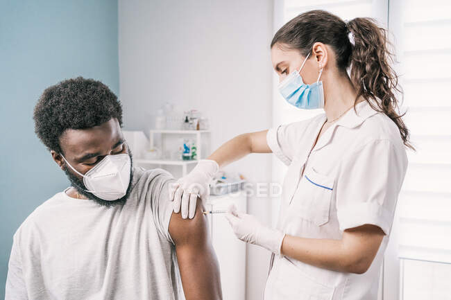 Специалистка-женщина в защитной форме, латексных перчатках и маске для лица, прививающая больного афроамериканца в клинике во время вспышки коронавируса — стоковое фото