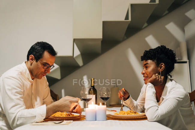 Vue latérale du couple multiracial heureux assis à table avec des bougies et du vin tout en mangeant de délicieux plats lors d'un dîner romantique à la maison — Photo de stock