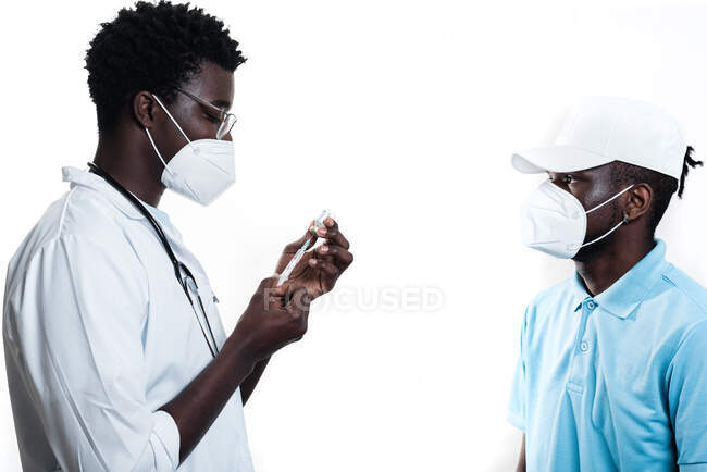 Médico étnico preenchendo seringa de frasco com vacina preparando-se para vacinar o paciente afro-americano masculino em fundo branco em uma clínica durante o surto de coronavírus — Fotografia de Stock