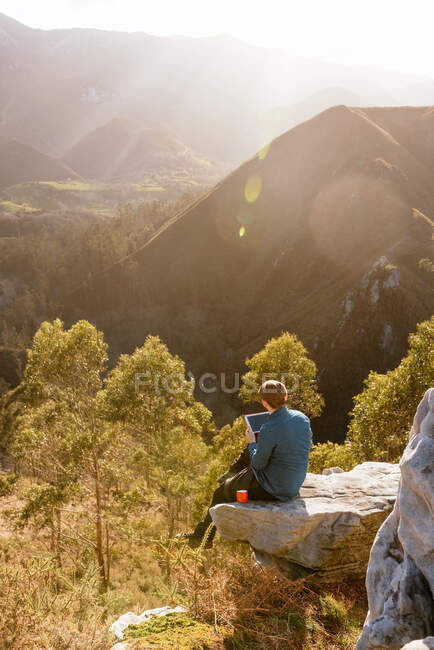 Tranquil male explorer with tablet sitting on hill in mountainous terrain at sunset — Stock Photo