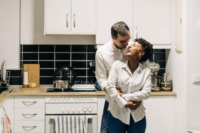 Uomo deliziato abbracciando sorridente donna nera da dietro mentre in piedi in cucina e guardandosi l'un l'altro — Foto stock