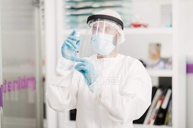 Вид збоку розмитого невідомого лікаря в захисній масці для обличчя та латексні рукавички з флаконом коронавірусної вакцини та шприцом, що стоїть у лікарняній кімнаті — стокове фото