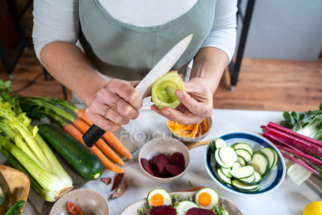 Von oben bis zur Unkenntlichkeit schneidet eine Person reife Avocado, während sie vegetarisches Essen mit verschiedenen Gemüsesorten zubereitet — Stockfoto