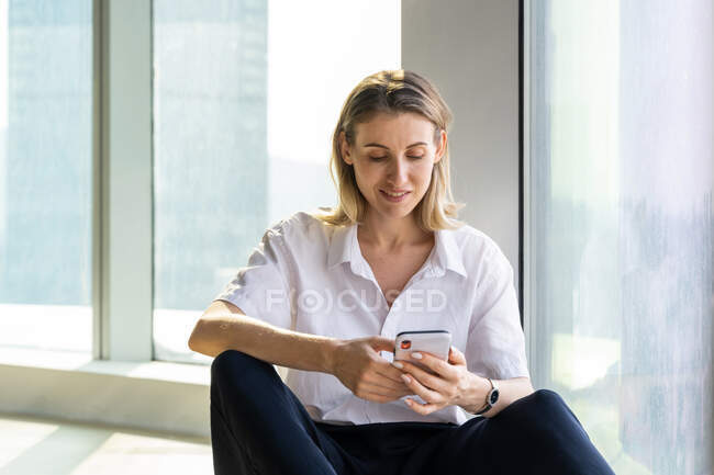 Delight young woman sitting in empty office with big window browsing on the mobile phone — Stock Photo