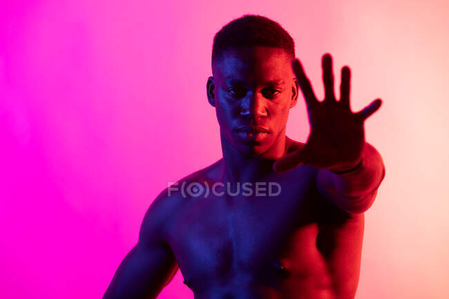 Серьезный молодой афроамериканец с обнаженным туловищем смотрит на камеру на розовом фоне в неоновой студии — стоковое фото
