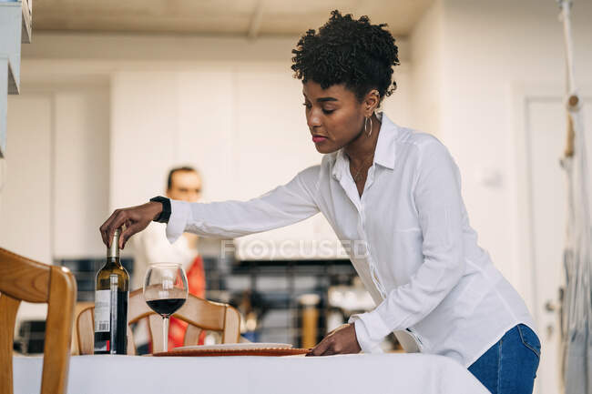 Вид сбоку на черную женщину, подающую красное вино на столе во время подготовки к романтическому ужину с парнем дома — стоковое фото