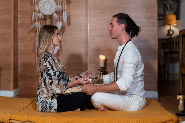 Vista lateral del maestro zen con la pareja femenina sentada con las piernas cruzadas y los ojos cerrados durante la práctica espiritual - foto de stock