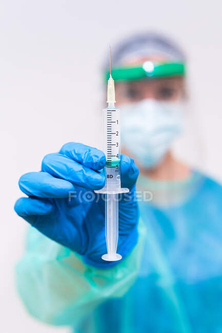Médecin flou méconnaissable dans un masque protecteur et gants en latex avec flacon de vaccin contre le coronavirus et seringue montrant à la caméra alors qu'il se tenait debout dans la chambre d'hôpital — Photo de stock