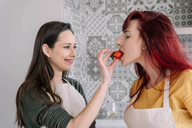 Молодая улыбающаяся женщина кормит гомосексуального партнера свежей клубникой против декоративной стены в доме — стоковое фото