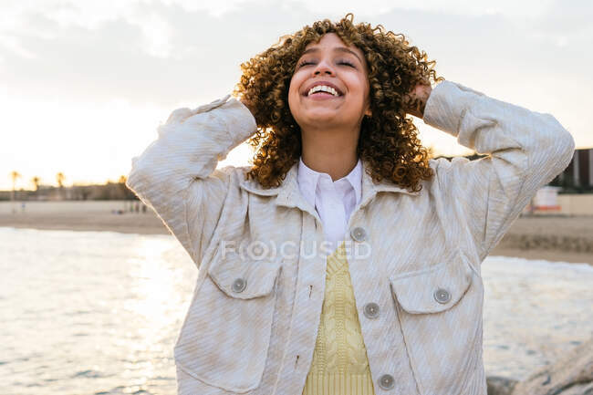Alto ángulo de alegre hembra afroamericana de pie con los brazos extendidos en las rocas en la orilla del mar y disfrutando de la libertad al atardecer - foto de stock