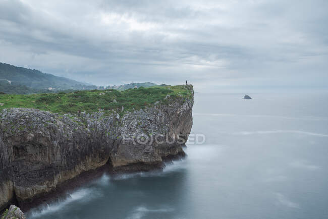 Silueta de persona parada al borde del acantilado rocoso cerca del mar en la costa de Ribadesella en día nublado en Asturias - foto de stock