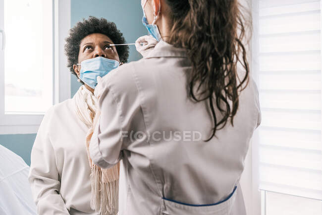 Especialista médica femenina en uniforme protector, guantes de látex y mascarilla facial haciendo prueba de coronavirus nasal en paciente madura afroamericana en clínica durante brote de virus - foto de stock