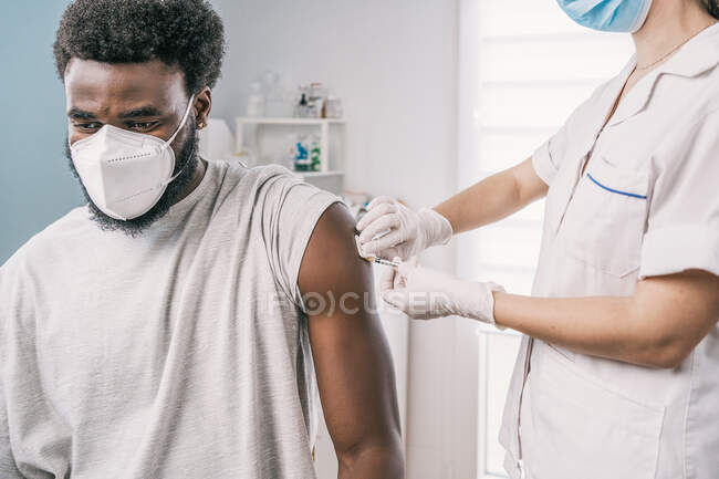 Ritagliato specialista medico femminile irriconoscibile in uniforme protettiva, guanti in lattice e maschera facciale vaccinare paziente uomo afroamericano in clinica durante l'epidemia di coronavirus — Foto stock