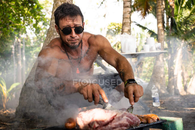 Shirtless adult male with sunglasses and necklace preparing tasty meat and sausages on grill on sunny day in nature — Stock Photo