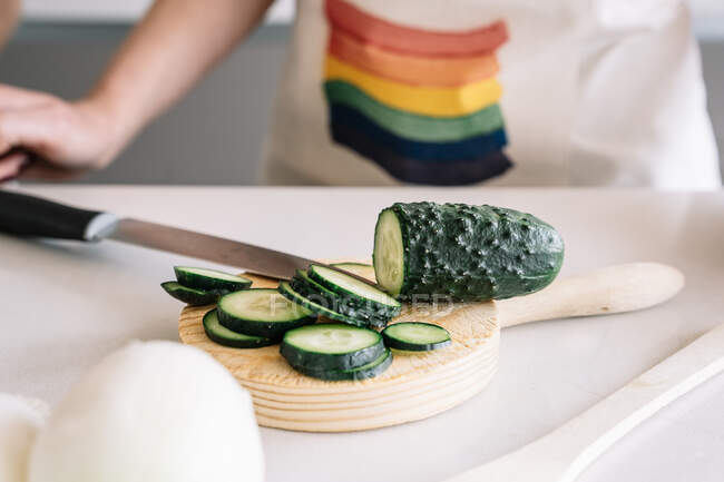 Crop unrecognizable homosexual female cutting fresh cucumber with knife while preparing healthy food at kitchen table with glass of wine — Stock Photo