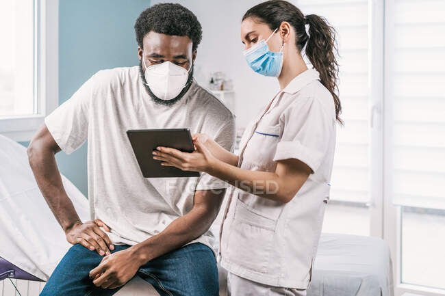 Jovem médica de uniforme médico e estetoscópio vestindo máscara facial falando e mostrando resultado em tablet para paciente homem afro-americano durante consulta na clínica — Fotografia de Stock