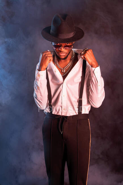 Young masculine ethnic male model in hat and trousers standing while looking at camera on black background with smoke — Stock Photo