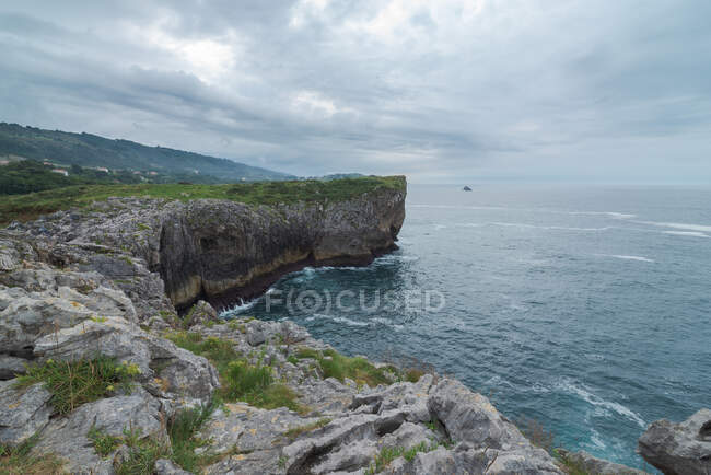 Impresionante vista del áspero acantilado rocoso cerca del tranquilo mar en la costa de Ribadesella bajo un cielo gris en Asturias - foto de stock