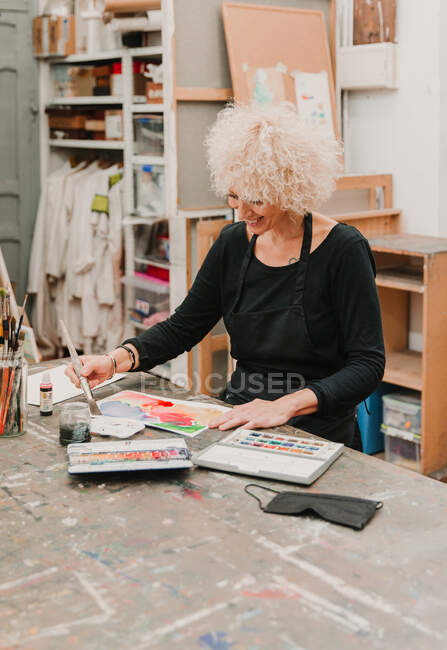 Focused female artist sitting at table and painting with watercolors on paper while working in creative workshop — Stock Photo
