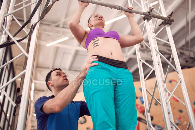 De baixo da fêmea fazendo pull ups na barra horizontal com a ajuda do instrutor enquanto treina no moderno centro de escalada — Fotografia de Stock