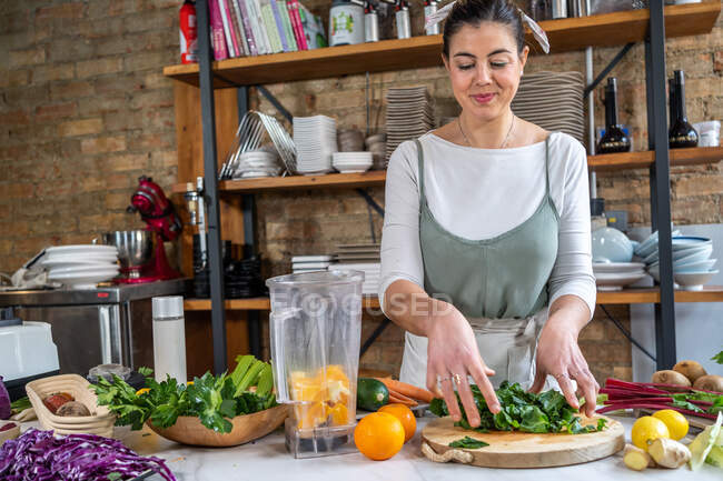 Жінка кладе свіже листя чаші блендера з апельсиновими скибочками в домашню кухню — стокове фото