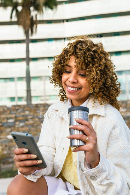 Femme afro-américaine positive avec une messagerie café à emporter sur les médias sociaux via smartphone dans la rue de la ville — Photo de stock