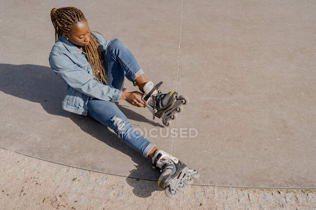 From above of African American female in denim outfit putting on rollerblades on sunny day in skate park — Stock Photo