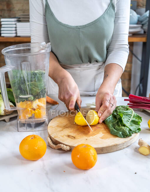 Corte anônimo fêmea corte limão suculento maduro com faca entre folhas de acelga e tigela de liquidificador na cozinha — Fotografia de Stock