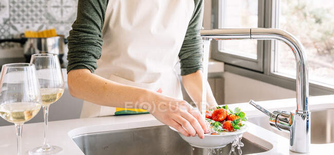 Cultiver jeune femelle dans le tablier laver les fraises fraîches sous l'eau liquide du robinet dans la cuisine maison — Photo de stock