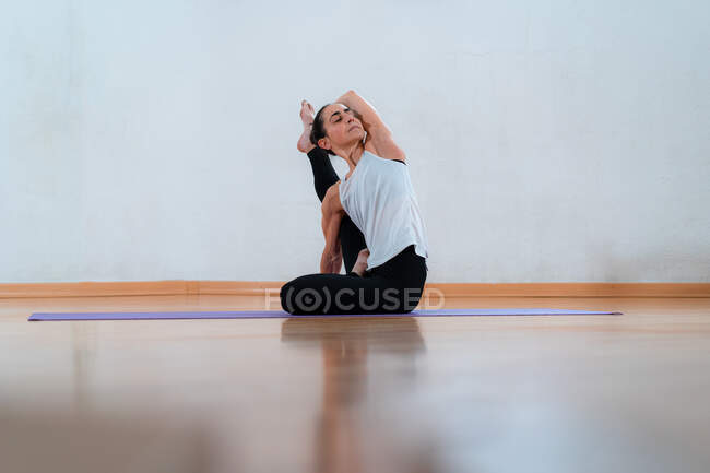 Уровень грунда женщины средних лет с закрытыми глазами в спортивной одежде, сидящей на коврике для йоги и растягивающей ногу в комнате — стоковое фото