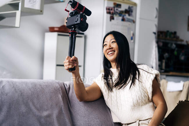 Lächelnde ethnische Vloggerin nimmt Video auf Fotokamera auf, während sie auf der Couch im Wohnzimmer sitzt — Stockfoto