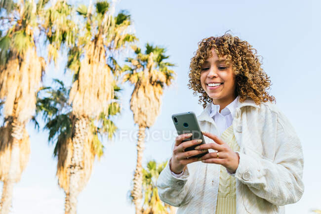 Niedriger Winkel einer fröhlichen afroamerikanischen Frau, die in einem exotischen Park mit Palmen bei Sonnenuntergang ihr Handy surft — Stockfoto