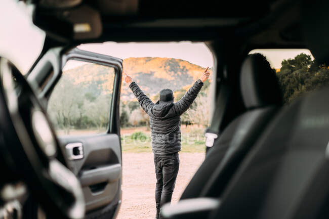 Vista trasera a través del interior de un coche de un hombre contemplando el paisaje montañoso al amanecer con los brazos levantados como signo de libertad - foto de stock