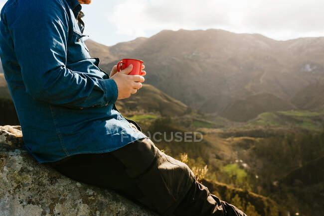 Vista lateral de un caminante masculino irreconocible recortado sentado en la colina con una taza de metal de bebida caliente y admirando un paisaje increíble de terreno montañoso en un día soleado - foto de stock