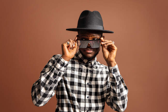 Молодой крутой афроамериканец в клетчатой рубашке, шляпе и солнцезащитных очках, стоя на коричневом фоне — стоковое фото