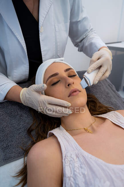 Crop cosmetologo irriconoscibile facendo peeling viso ultrasonico per donna rilassata sdraiata sul tavolo medico nella moderna clinica di bellezza — Foto stock