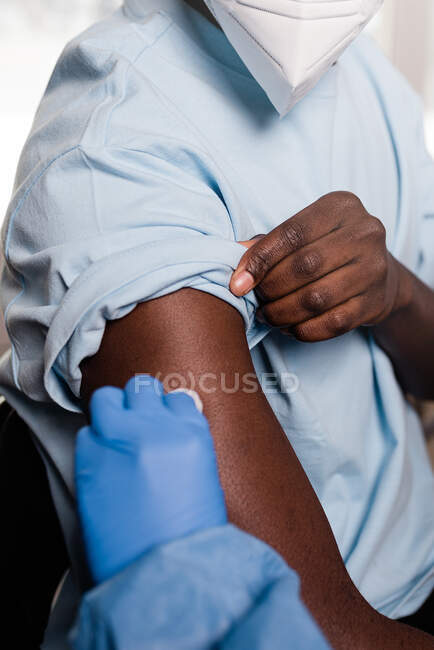 Spécialiste médical méconnaissable en uniforme de protection et gants en latex désinfectant le bras avec du coton avec de l'alcool pour vacciner le patient afro-américain masculin en clinique pendant l'épidémie de coronavirus — Photo de stock