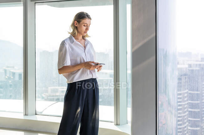 Mujer joven solitaria y sin emociones de pie en una oficina vacía con una gran ventana navegando en el teléfono móvil - foto de stock