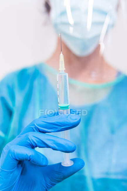Blurred unrecognizable medic in protective face shield mask and latex gloves with vial of coronavirus vaccine and syringe showing to camera while standing in hospital room — Stock Photo