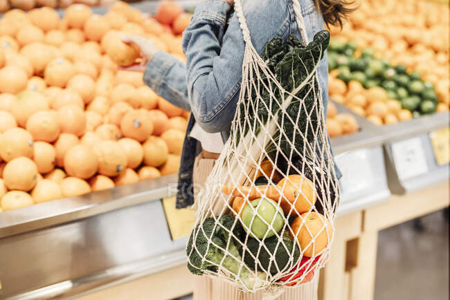 Vista lateral da colheita comprador feminino irreconhecível em pé no supermercado com saco de malha eco friendly cheio de frutas e legumes maduros — Fotografia de Stock