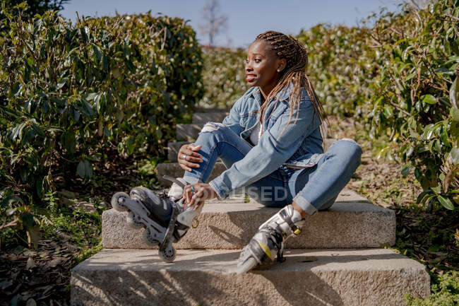 Улыбающаяся афроамериканка надевает ролики, сидя на лестнице в летнем парке и отворачиваясь — стоковое фото